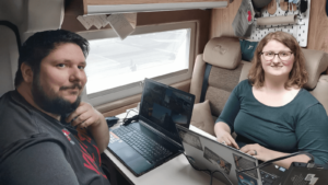 Zwei Personen arbeiten an Laptops in einem gemütlichen Campervan, das als mobiles Büro eingerichtet ist. Sie lächeln in die Kamera, während sie an einem Tisch sitzen, der für das ortsunabhängige Arbeiten ausgestattet ist.