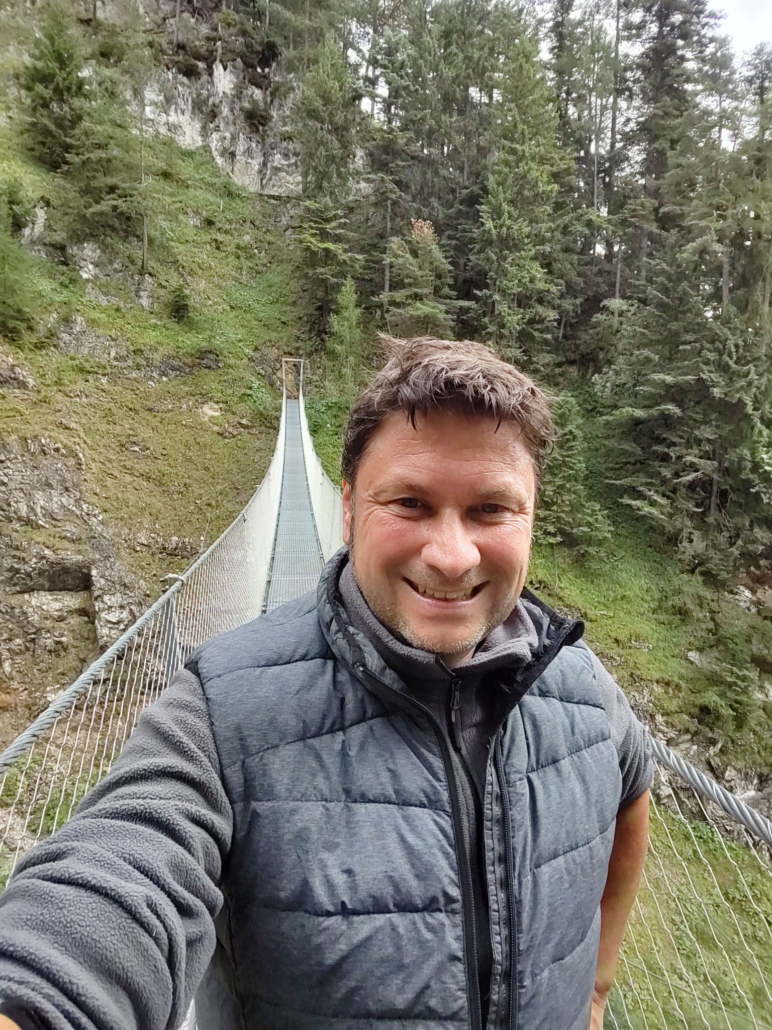 Hiker taking selfie on mountain suspension bridge A man smiling on a suspension bridge surrounded by tall pine trees and rocky cliffs