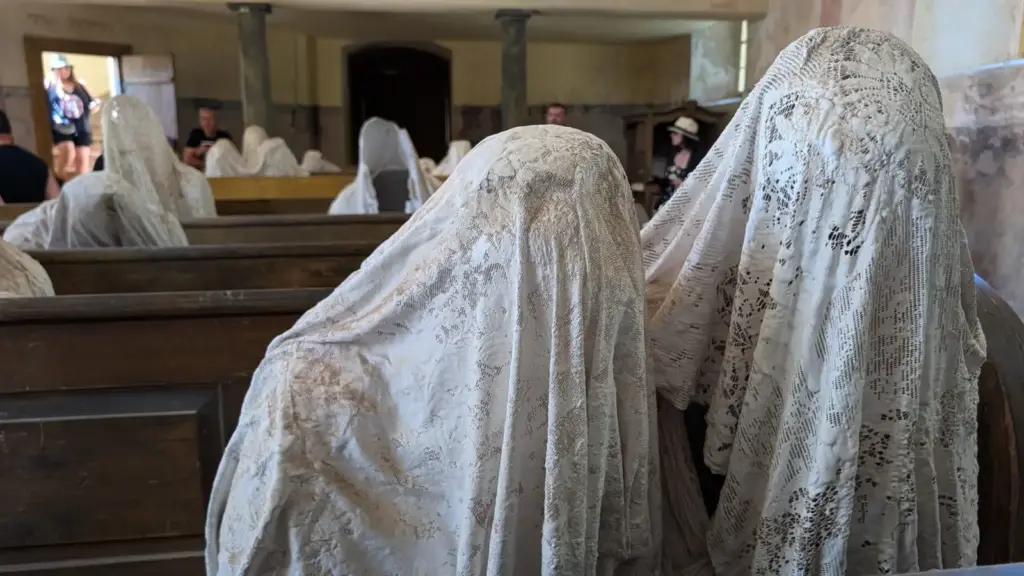 Nahaufnahme zweier mit historischen Spitzentüchern verhüllter Geisterfiguren in Kirchenbänken der Geisterkirche Lukova, im Hintergrund Besucher